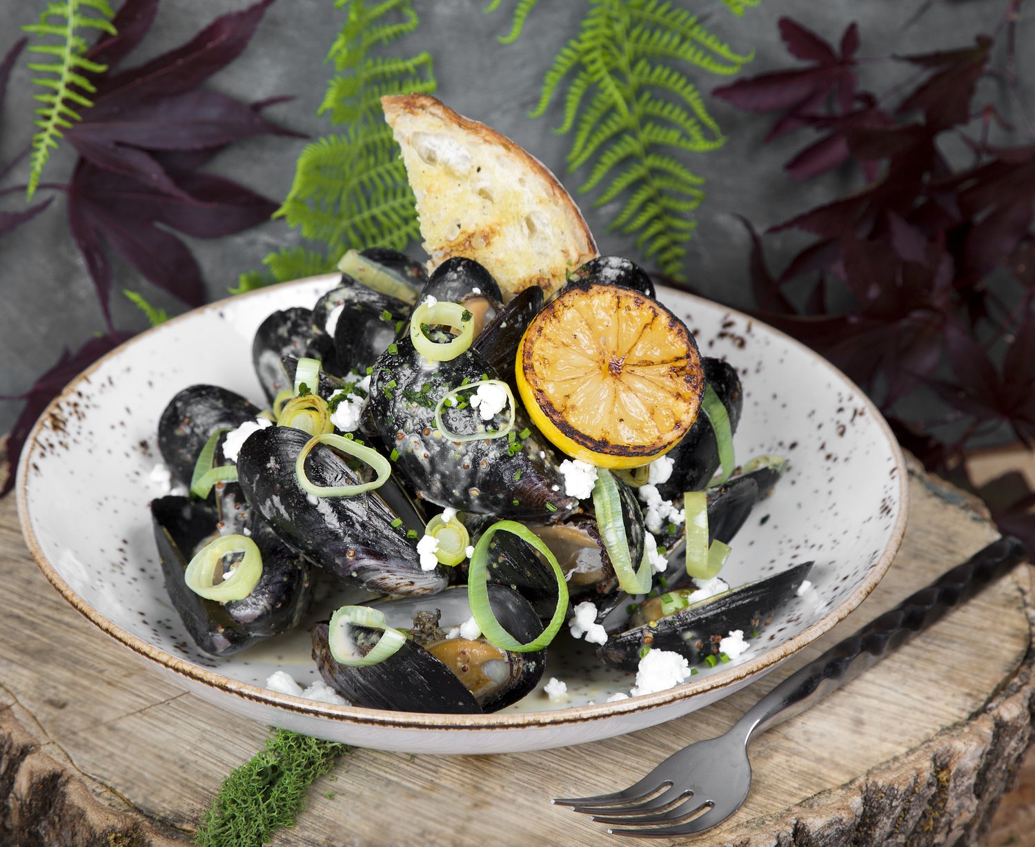 A bowl of mussels with grilled bread, greens and a charred lemon.