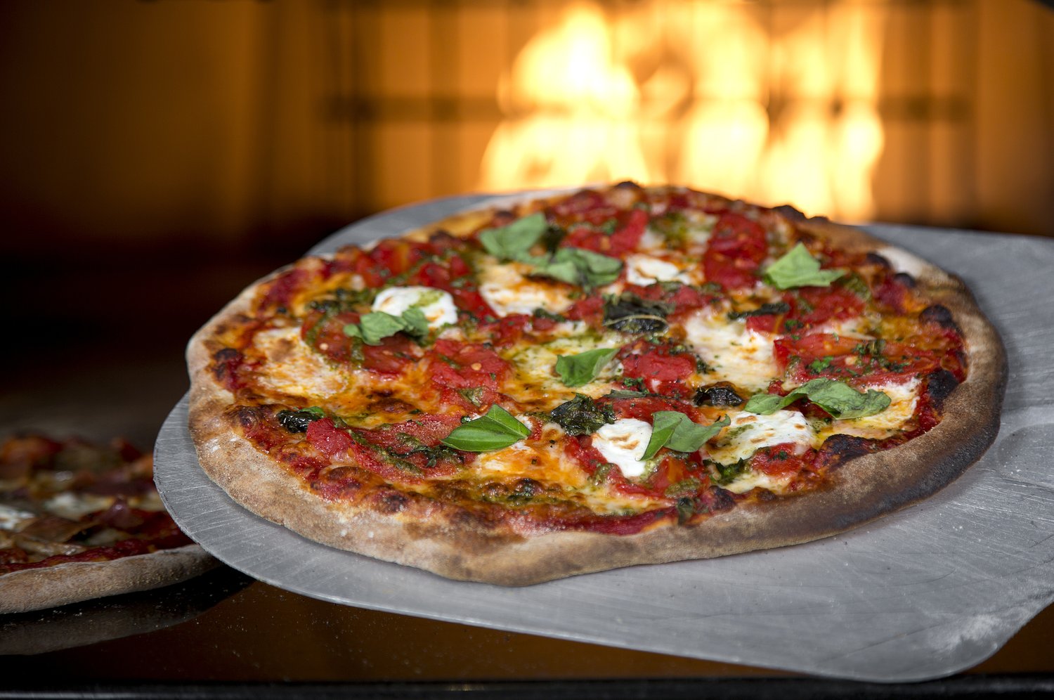 A wood-fired pizza on a stone, with the oven flames glowing behind it.