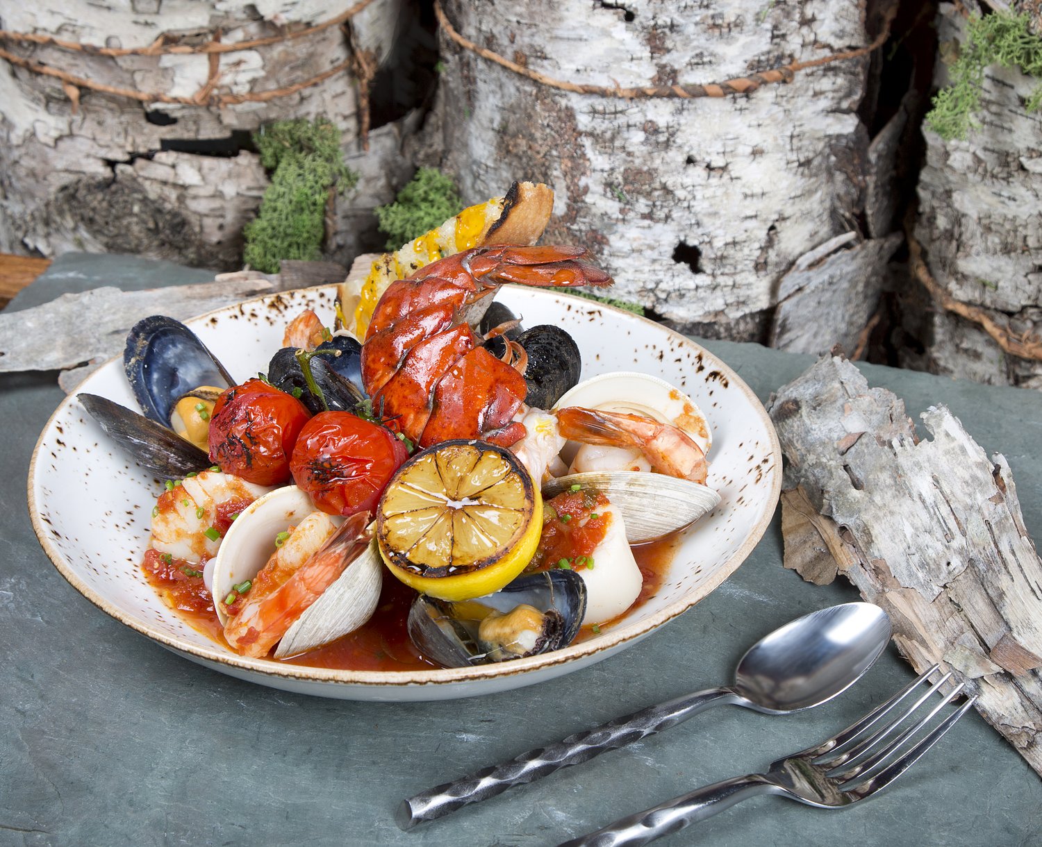 A wide bowl of shellfish stew with shrimp, mussels and lobster in a red broth, on a wood round.